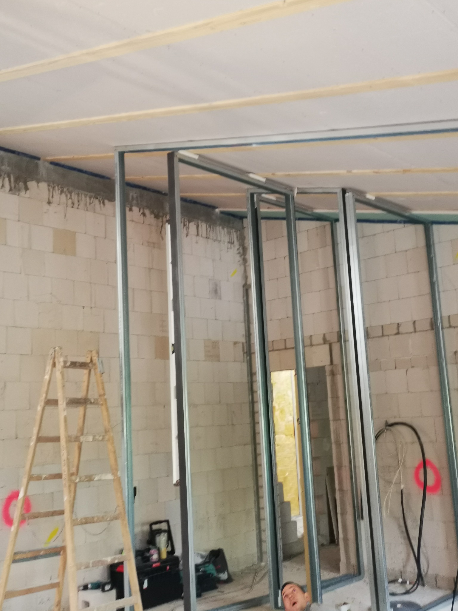 Konstrukcja szkieletowa z metalowych profili na tle ściany z betonu komórkowego, widoczna drewniana drabina i narzędzia, przygotowania do montażu płyt gipsowo-kartonowych na suficie.