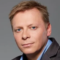 Portret mężczyzny z krótkimi blond włosami, w niebieskiej koszuli i ciemnej marynarce, na szarym tle.