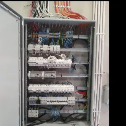 Otwarta skrzynka elektryczna z licznymi bezpiecznikami, przekaźnikami i przewodami w kolorach niebieskim, czarnym, czerwonym i pomarańczowym, umieszczona na ścianie z widocznymi rurami ochronnymi.