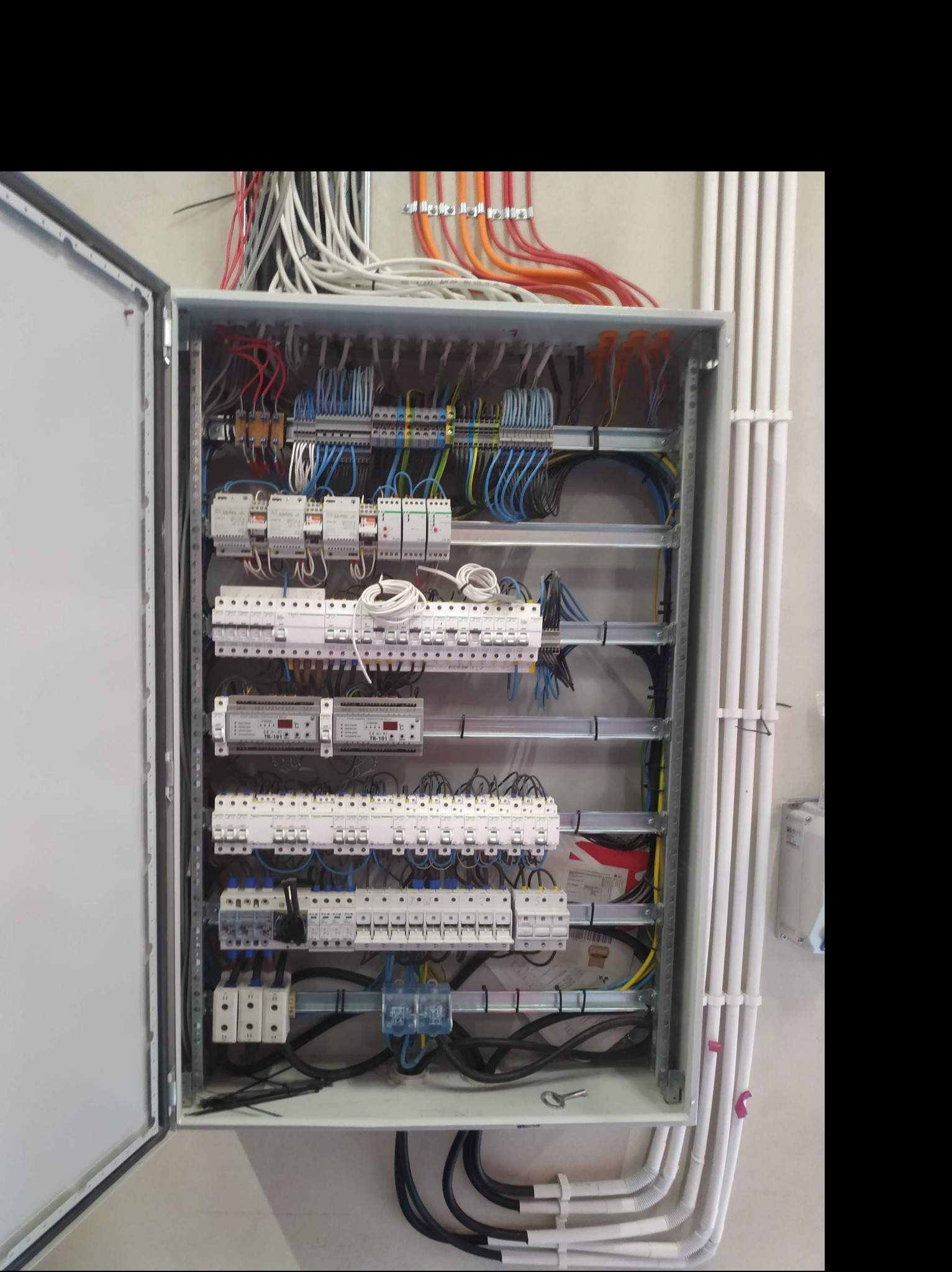 Otwarta skrzynka elektryczna z licznymi bezpiecznikami, przekaźnikami i przewodami w kolorach niebieskim, czarnym, czerwonym i pomarańczowym, umieszczona na ścianie z widocznymi rurami ochronnymi.