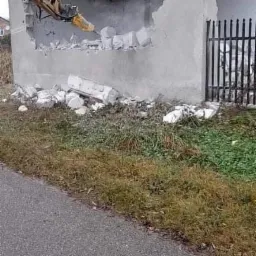 Żółta koparka z młotem pneumatycznym wyburza betonową ścianę budynku; gruz leży na ziemi, widoczne metalowe ogrodzenie i fragment drogi.