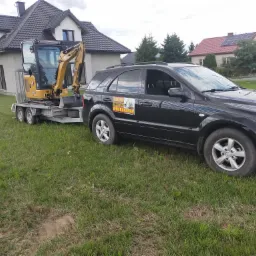 Czarny SUV zaparkowany na trawniku przed domem, obok przyczepa z małą koparką CAT 301.8. Na drzwiach samochodu naklejka reklamowa firmy oferującej usługi minikoparką.