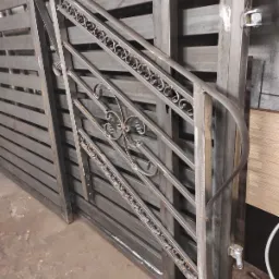 Surowa, spawana, metalowa konstrukcja bramy z ozdobnymi elementami, widoczne ślady spawania i niedoskonałości wykończenia.