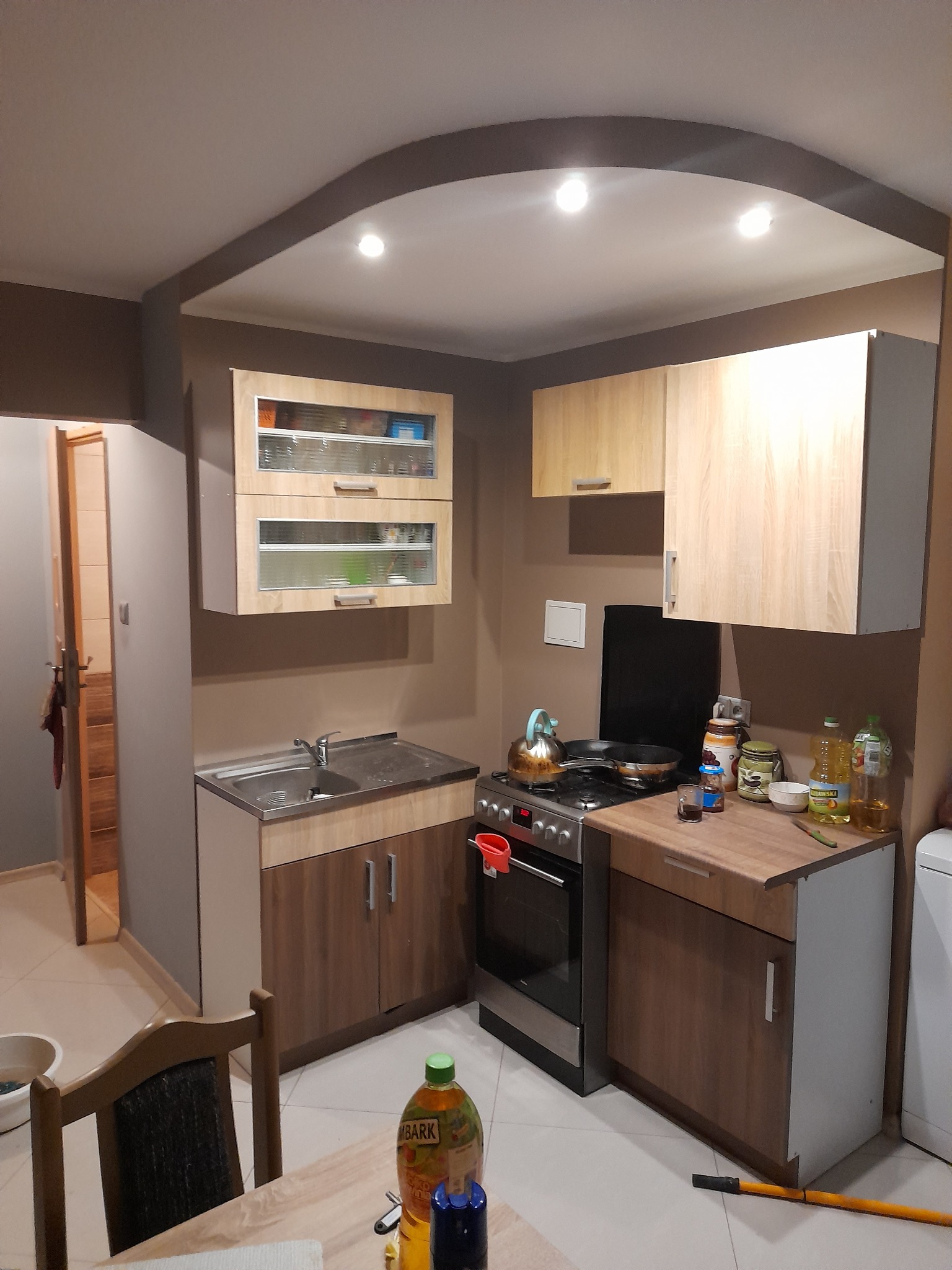 Umeblowana kuchnia w brązowo-beżowych tonacjach z szafkami wiszącymi z przeszklonymi frontami, metalowym zlewem i kuchenką gazową z czajnikiem.