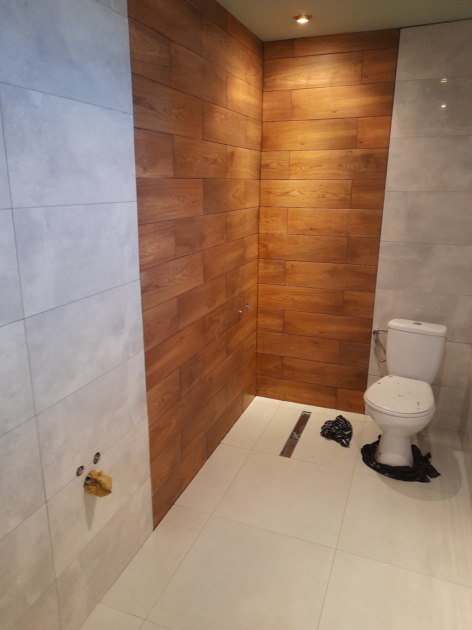 Wykończona łazienka z drewnopodobnymi płytkami na ścianie, jasnymi płytkami podłogowymi i wiszącą muszlą klozetową, widoczny odpływ liniowy i otwory w ścianie zabezpieczone folią.