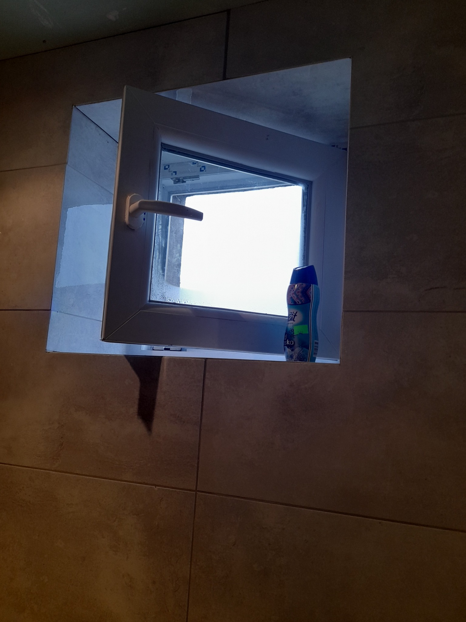 Otwarty biały okno PCV w łazience wyłożonej brązowymi płytkami, na parapecie stoi niebieski odświeżacz powietrza.