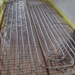 Białe rurki ogrzewania podłogowego ułożone na folii z czerwoną kratką, zabezpieczone żółtą folią przy ścianach, widok z góry.