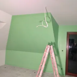 Pokój na poddaszu z pomalowanymi na zielono ścianami, drewnianą ramą drzwi i rozłożoną drabiną, widoczne wystające przewody elektryczne.