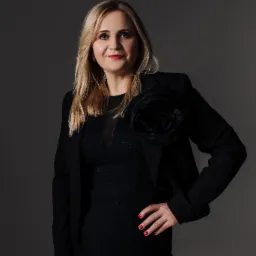 Portret kobiety w czarnej sukience i marynarce z ozdobną różą, prezentującej profesjonalny wizerunek na szarym tle.