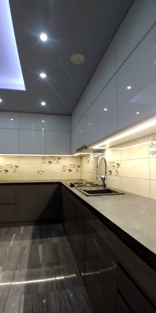 Nowoczesna kuchnia z ciemnymi, lakierowanymi frontami, jasnymi górnymi szafkami i blatem w odcieniach szarości, oświetlona punktowymi lampami sufitowymi i listwą LED pod szafkami, z płytkami...