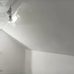 Świeżo pomalowane, jasne poddasze ze skosami. Na suficie widoczny fragment zabezpieczonej lampy. Ściany i sufit w jednolitej, matowej bieli.