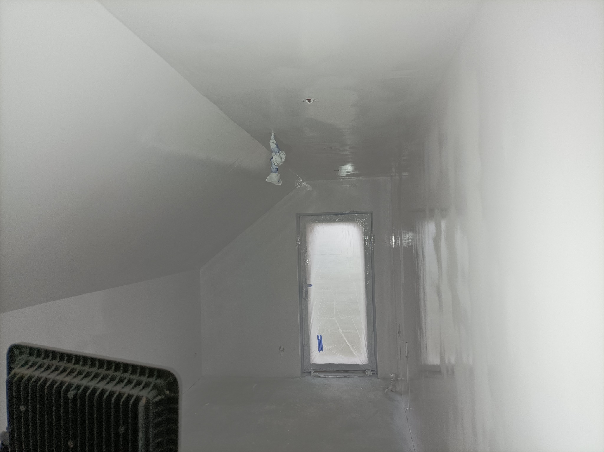Poddasze po świeżym malowaniu na biało, okno zabezpieczone folią, widoczny grzejnik elektryczny, z sufitu zwisa zaklejona taśmą malarską lampa.