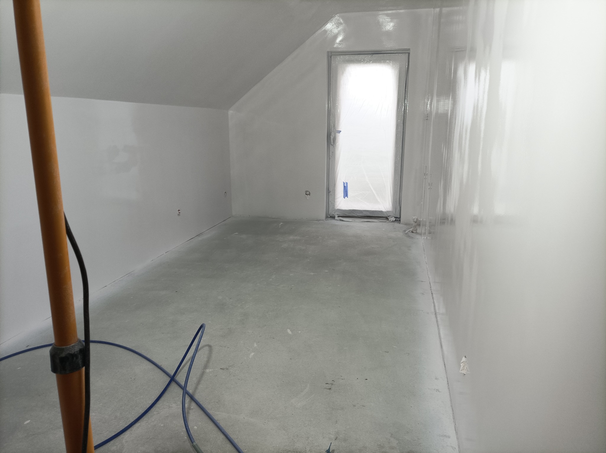 Poddasze po świeżym, jednolitym pomalowaniu na biało, z zabezpieczonym folią oknem dachowym i widocznym przewodem od agregatu malarskiego.