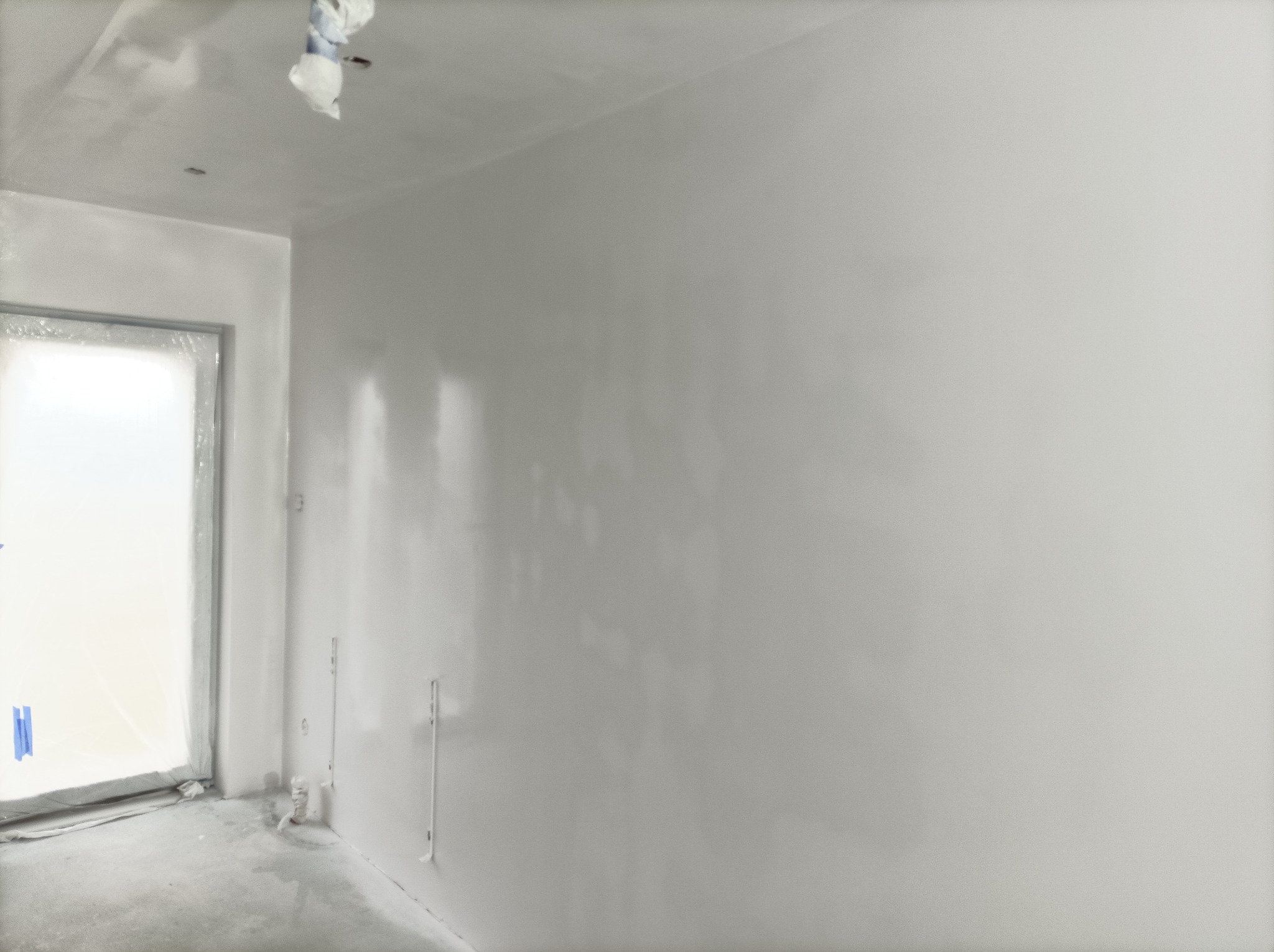 Świeżo pomalowane na biało ściany i sufit w pokoju, z zabezpieczonym folią oknem i widocznymi puszkami elektrycznymi, ukazujące etap prac malarskich.