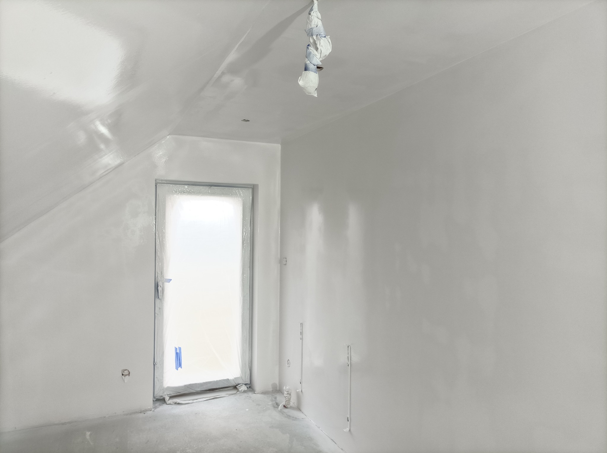 Poddasze po świeżym malowaniu na biało, zabezpieczone folią okno, widoczne ślady po szpachlowaniu na suficie.
