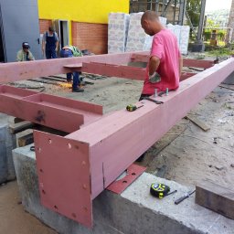 R&R Development - Pracownicy budowlani montują stalową konstrukcję nośną, spawanie elementów na zewnątrz budynku, widoczne iskry i narzędzia pomiarowe.