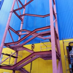 R&R Development - Montaż stalowej konstrukcji na tle niebiesko-żółtej ściany budynku, widoczne rusztowania i pracownik w niebieskiej kurtce.