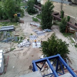 R&R Development - Widok z góry na plac budowy z porozrzucanymi elementami konstrukcyjnymi, w oddali widoczni pracownicy przy montażu, a w tle budynki fabryczne i rury ciepłownicze.