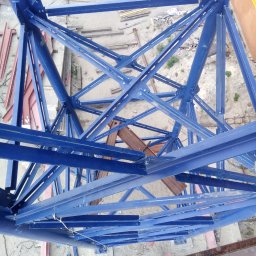 R&R Development - Konstrukcja stalowa w kolorze niebieskim, widok z dołu na siatkę belek i słupów, elementy montażowe na placu budowy widoczne w tle.