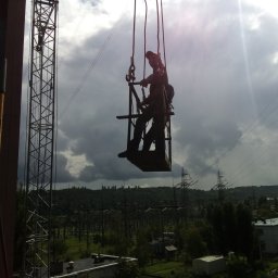R&R Development - Pracownik w uprzęży na platformie podwieszonej do dźwigu, wykonujący prace na wysokości przy słupach wysokiego napięcia na tle pochmurnego nieba.
