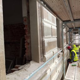 R&R Development - Pracownik w kasku i kamizelce na rusztowaniu, w trakcie renowacji elewacji budynku z elementami dekoracyjnymi, widoczne odsłonięte cegły i narzędzia.