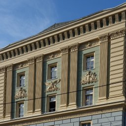 R&R Development - Elewacja budynku z kolumnami, dekoracyjnymi płaskorzeźbami, oknami i listwami w odcieniach beżu, zieleni i szarości pod błękitnym niebem.