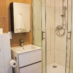 Nowoczesna łazienka z kabiną prysznicową z chromowanymi profilami, białą umywalką z szafką, lustrem z wiszącym ręcznikiem i brązowymi płytkami na ścianie, oświetlona punktowym światłem.