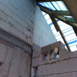 R&R Development - Wznoszenie ściany z bloczków betonu komórkowego, widoczny fragment drewnianej konstrukcji dachu zabezpieczonej folią, poziomica przymocowana do ściany.