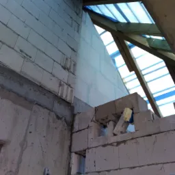 Wznoszenie ściany z bloczków betonu komórkowego, widoczny fragment drewnianej konstrukcji dachu zabezpieczonej folią, poziomica przymocowana do ściany.