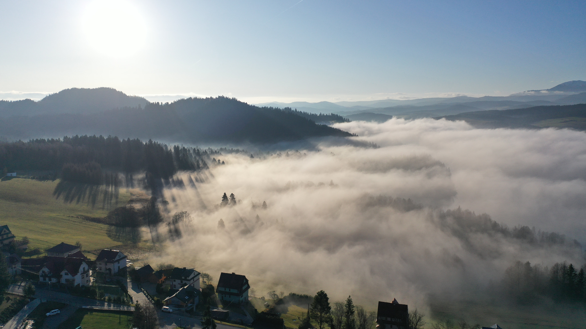 Panoramiczny widok z lotu ptaka na wieś spowitą gęstą mgłą, z zalesionymi wzgórzami w tle i delikatnym światłem słonecznym przebijającym się przez chmury.