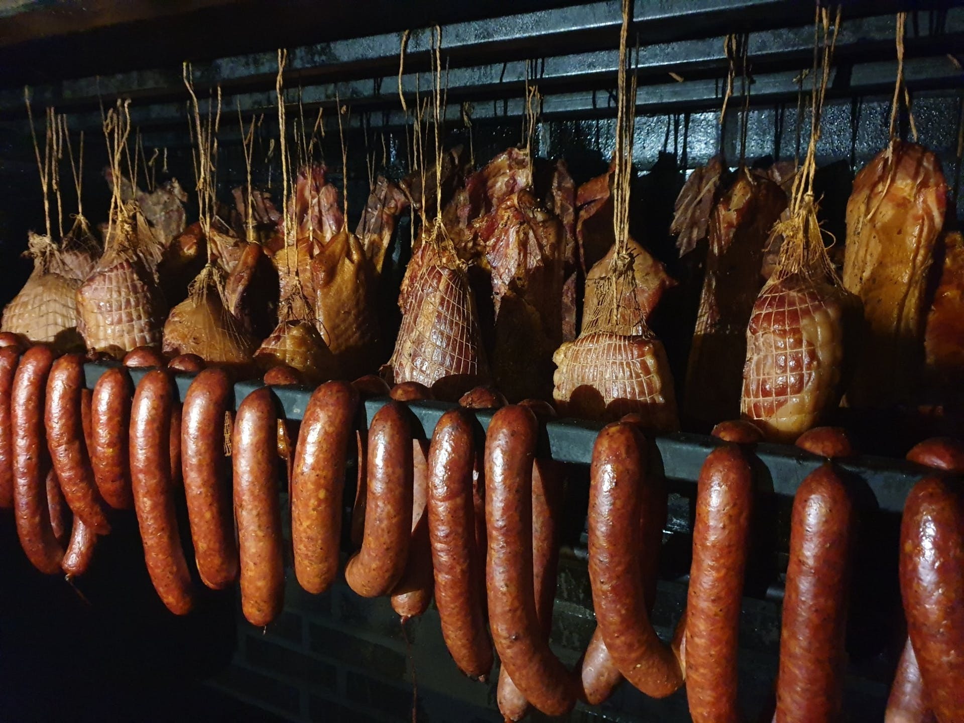 Wędzone kiełbasy i wędzonki wiszące w wędzarni, widoczne sznurki i siatki wędliniarskie, ciemne otoczenie.