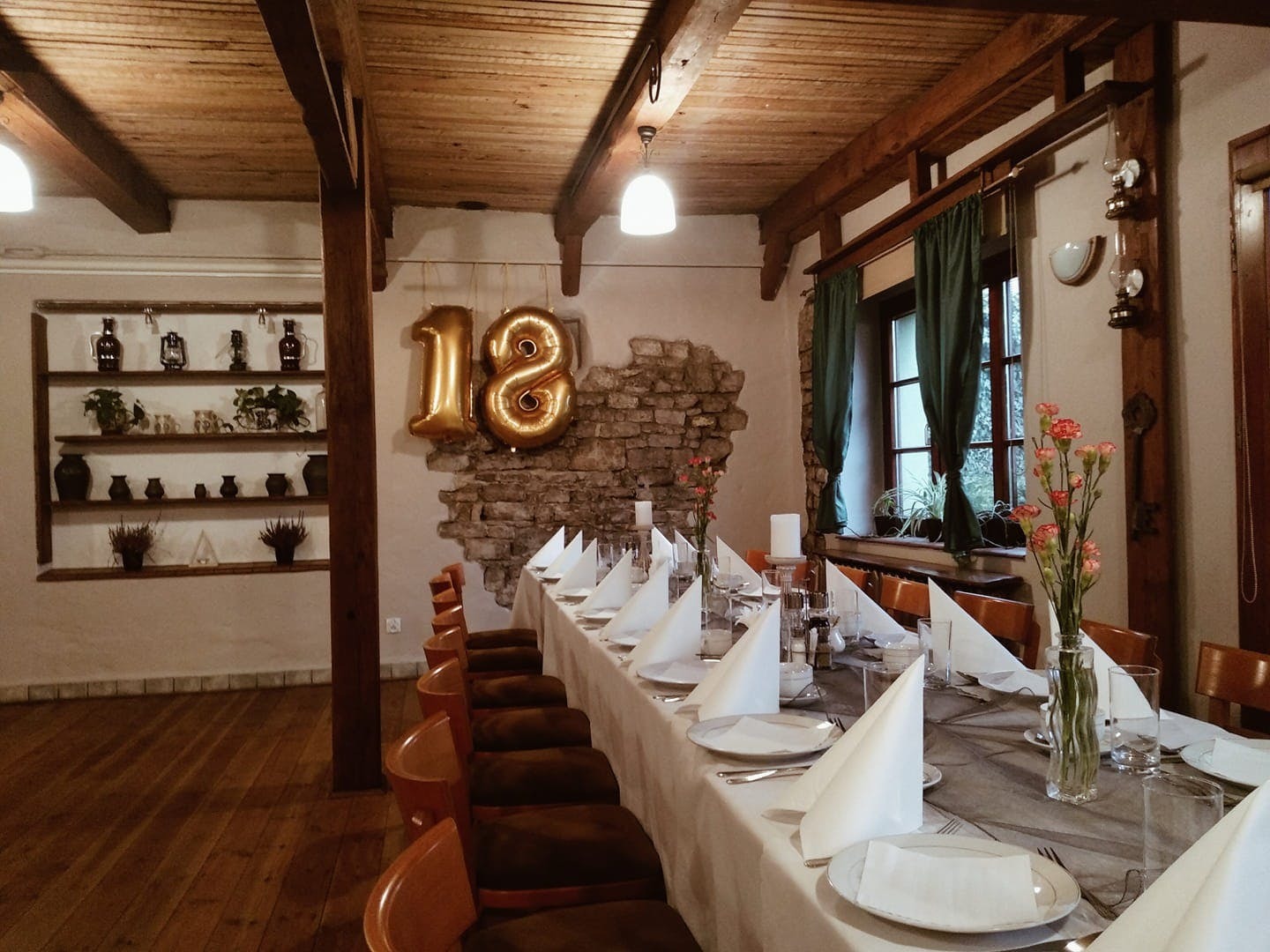 Uroczysta sala z długim stołem nakrytym białym obrusem, elegancko złożone serwetki, wazony z różowymi goździkami, złote balony w kształcie liczby 18, drewniany sufit z belkami, półka z dekoracjami...