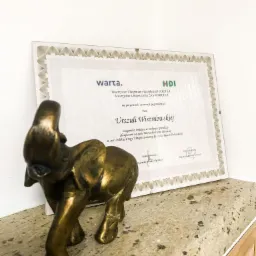 Statuetka słonia z brązu na tle oprawionego dyplomu z logo Warta i HDI, gratulującego Urszuli Wiszniewskiej osiągnięcia pierwszego miejsca w rankingu sprzedaży ubezpieczeń na życie.