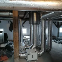 System rur wentylacyjnych w izolacji aluminiowej w pomieszczeniu z drewnianą konstrukcją stropu i kolumną nośną.