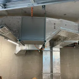 System kanałów wentylacyjnych z izolacją aluminiową i centralą w szarej obudowie, zamontowany pod sufitem w surowym wnętrzu budynku komercyjnego.