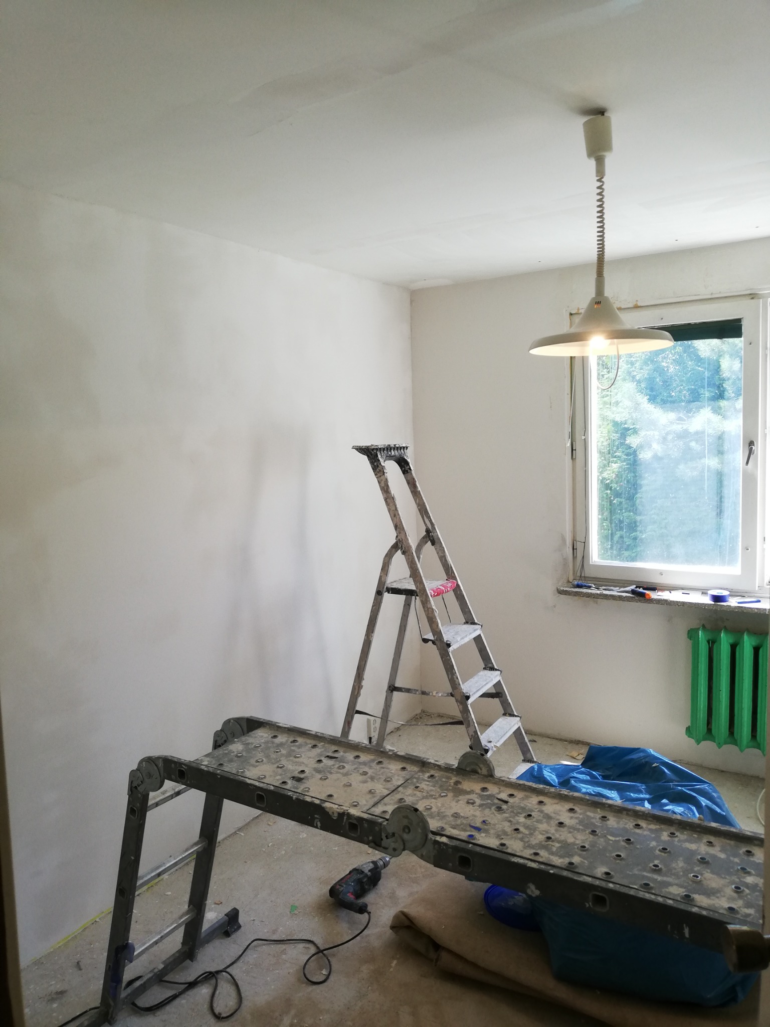 Pokój w trakcie odświeżania: świeżo wyszpachlowane ściany, drabina aluminiowa oparta o ścianę, platforma robocza z wiertarką na podłodze, okno z zielonym grzejnikiem.