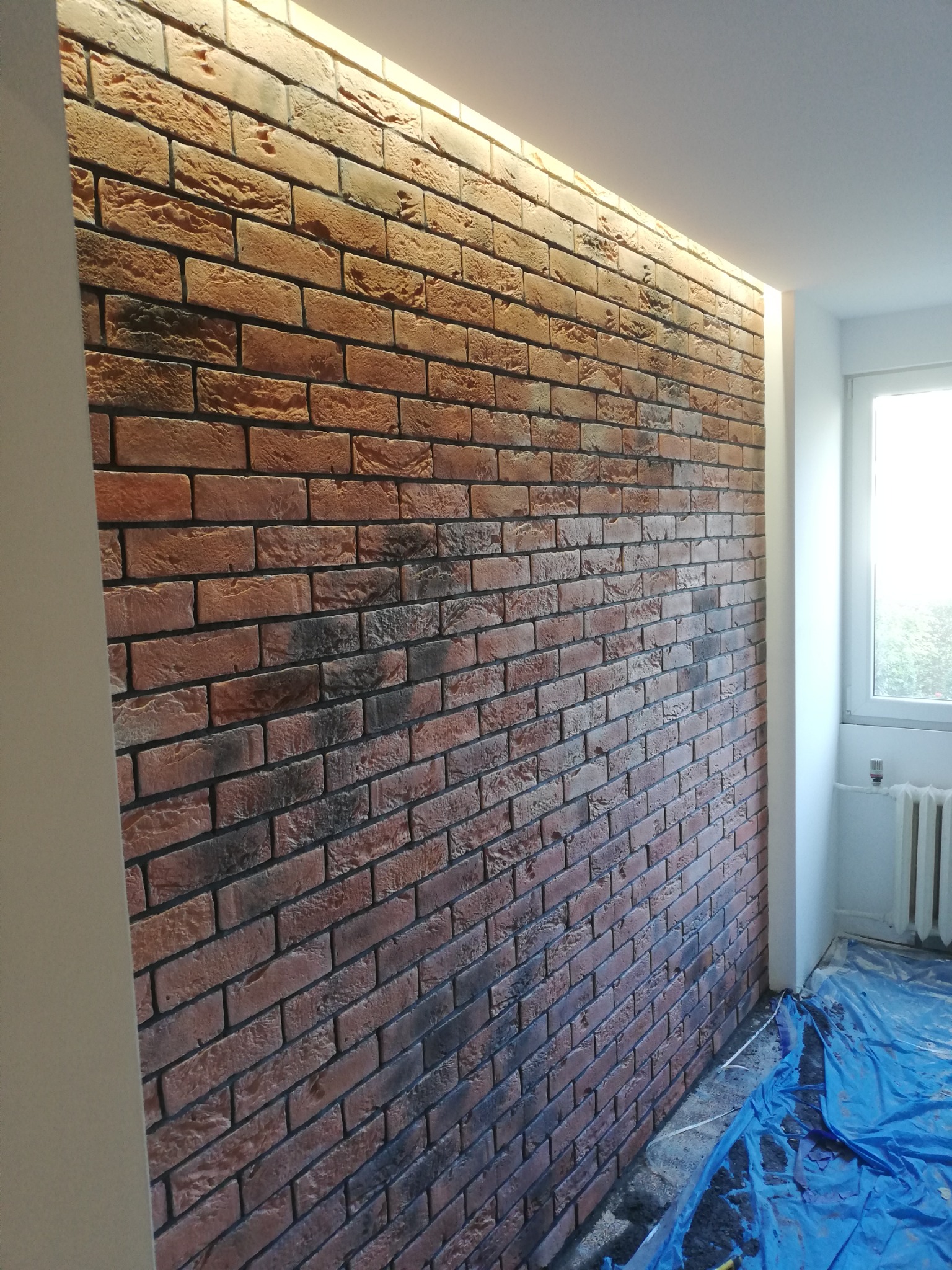 Ściana wyłożona dekoracyjną cegłą klinkierową z podświetleniem LED wzdłuż sufitu; zabezpieczona podłoga folią malarską.