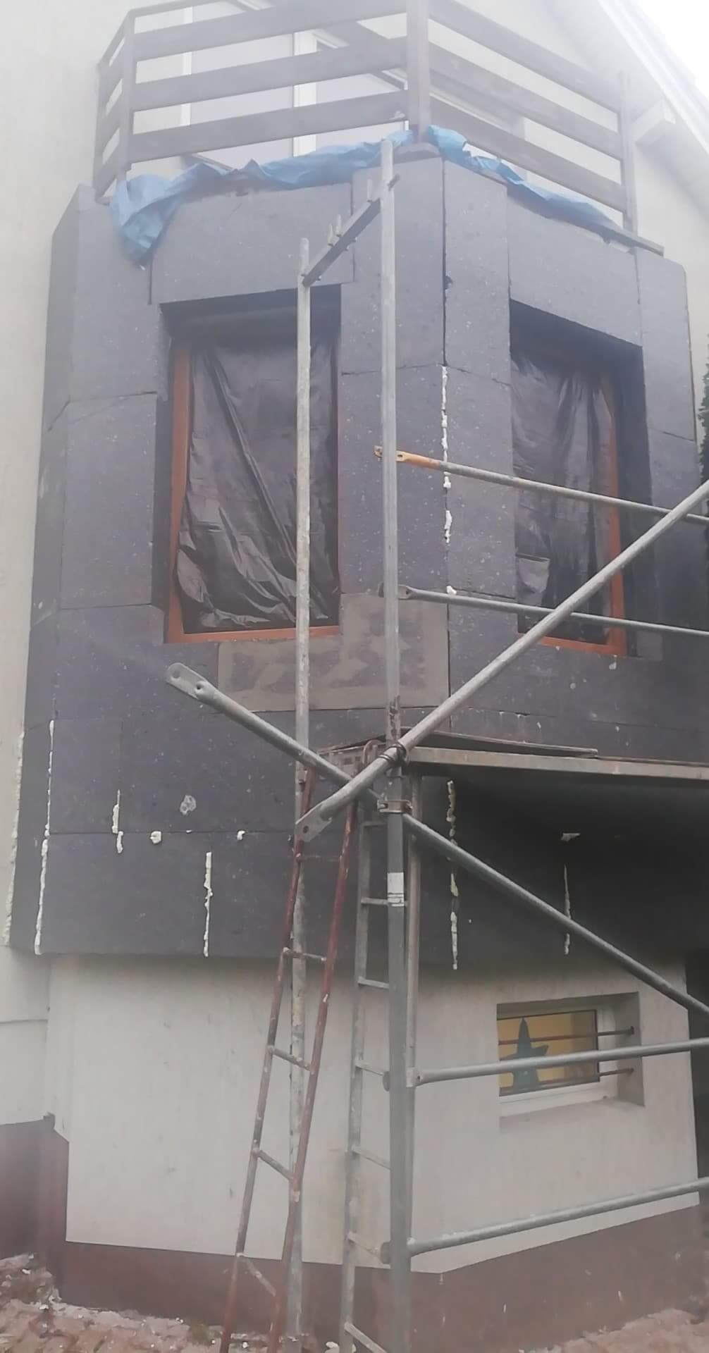 Ocieplanie elewacji budynku styropianem grafitowym, widoczne rusztowanie i okna zabezpieczone folią.