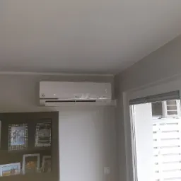 Biały klimatyzator LG zamontowany na szarej ścianie w pokoju, widoczny fragment okna z żaluzjami i ciemne meble z ramkami na zdjęcia.