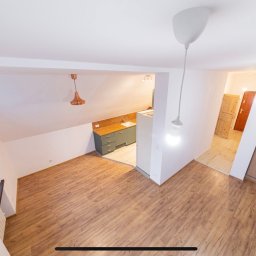 Stefan Kraft Spółka z Ograniczoną Odpowiedzialnością - Wnętrze mieszkania na poddaszu z drewnianą podłogą, widok na aneks kuchenny z szarymi szafkami i drewnianym blatem oraz przesuwne drzwi do szafy, oświetlone wiszącymi lampami bez kloszy.