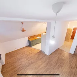 Wnętrze mieszkania na poddaszu z drewnianą podłogą, widok na aneks kuchenny z szarymi szafkami i drewnianym blatem oraz przesuwne drzwi do szafy, oświetlone wiszącymi lampami bez kloszy.