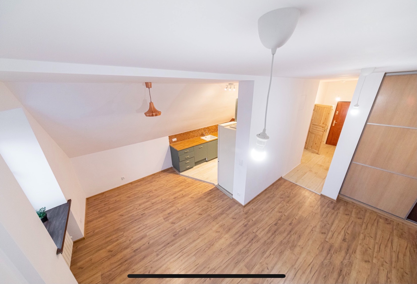 Wnętrze mieszkania na poddaszu z drewnianą podłogą, widok na aneks kuchenny z szarymi szafkami i drewnianym blatem oraz przesuwne drzwi do szafy, oświetlone wiszącymi lampami bez kloszy.