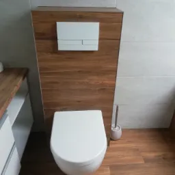 Nowoczesna toaleta podwieszana z zabudową z płyty drewnopodobnej, biały przycisk spłukiwania, szare płytki ścienne i drewnopodobne płytki podłogowe, szczotka do WC w rogu.