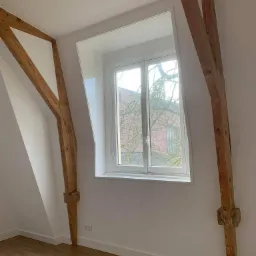 Poddasze po remoncie: Świeżo pomalowane białe ściany z widocznymi drewnianymi belkami konstrukcyjnymi, okno z widokiem na budynek i drzewo, parkiet na podłodze i gniazdka elektryczne przy listwie...