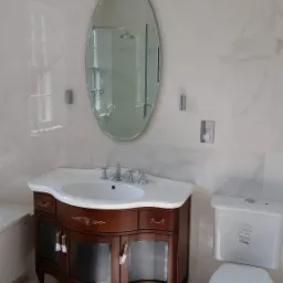 Klasyczna łazienka z umywalką na drewnianej szafce z przeszklonymi drzwiczkami, owalne lustro, biała armatura i marmurowe płytki ścienne.