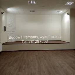 Pusta sala z podwyższonym podestem, wykończona panelami podłogowymi w odcieniach brązu, biała ściana z fragmentem dekoracyjnej cegły i oświetleniem sufitowym.