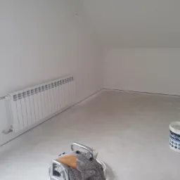 Świeżo pomalowane na biało poddasze z grzejnikiem, wiaderkiem z farbą i ubraniem na metalowym wózku.