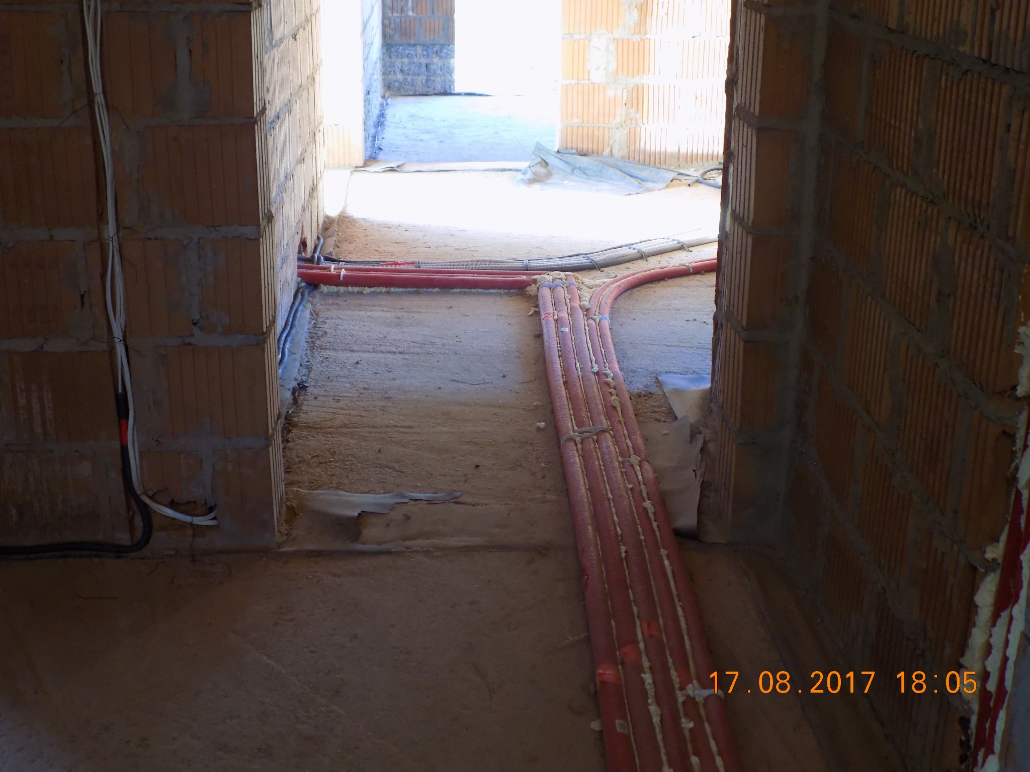 Wnętrze w trakcie budowy z widocznymi rurami instalacji grzewczej w czerwonej izolacji, ułożonymi na podłodze w przejściu między pomieszczeniami, oraz przewodami elektrycznymi przy ścianie z cegły...