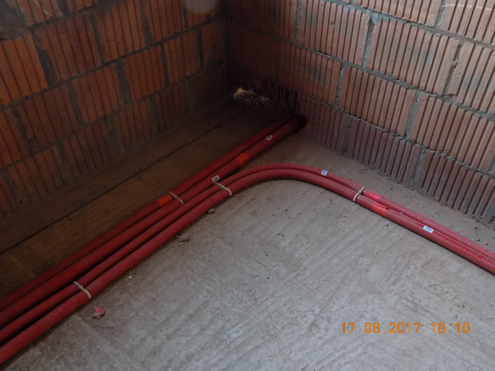 Czerwone rury ogrzewania podłogowego ułożone na szarym podłożu w rogu pomieszczenia z ceglanymi ścianami w trakcie instalacji.