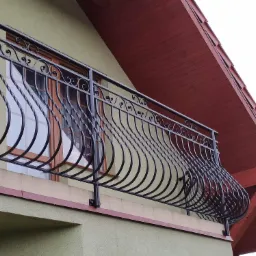 Czarna, ozdobna balustrada balkonowa z giętych elementów, zamontowana na balkonie domu z oliwkową elewacją i czerwonym dachem.